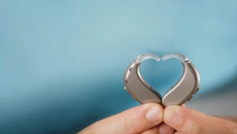 Two behind-the-ear hearing aids held in the shape of a heart with a blue sky background.