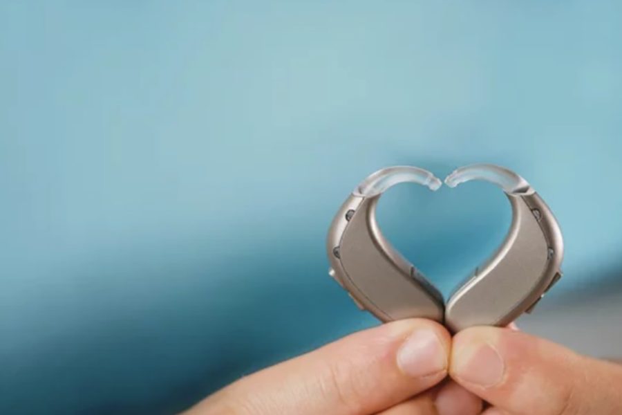 Two behind-the-ear hearing aids held in the shape of a heart with a blue sky background.
