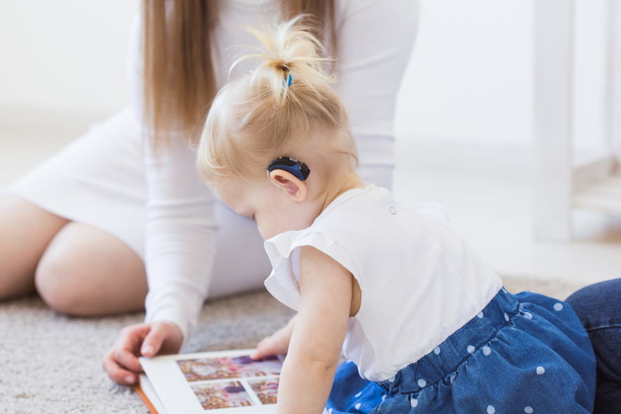 A child with a hearing aid is reading a book on the ground with her speech-language pathologist.