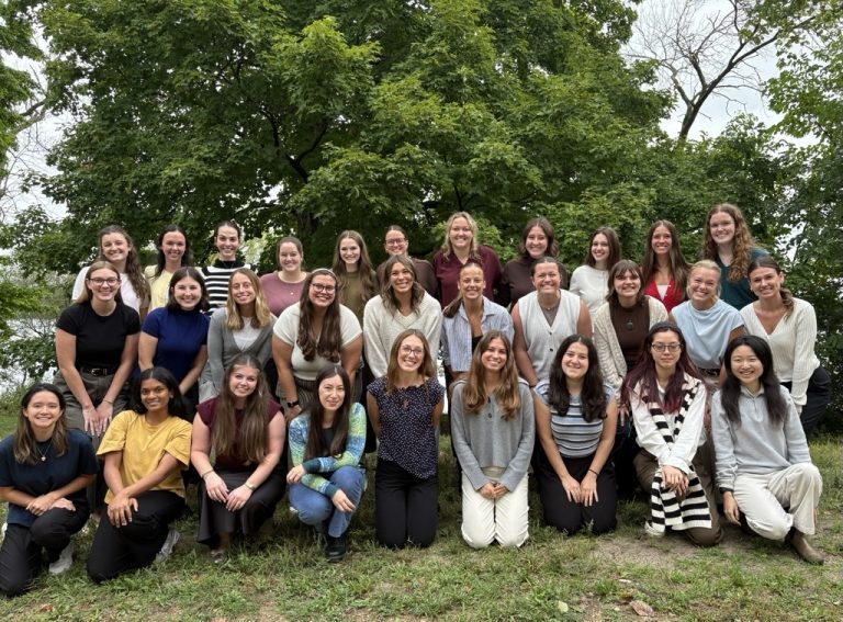 Group of first year speech graduate students gathered together outside of Goodnight Hall, with Lake Mendota in the background.